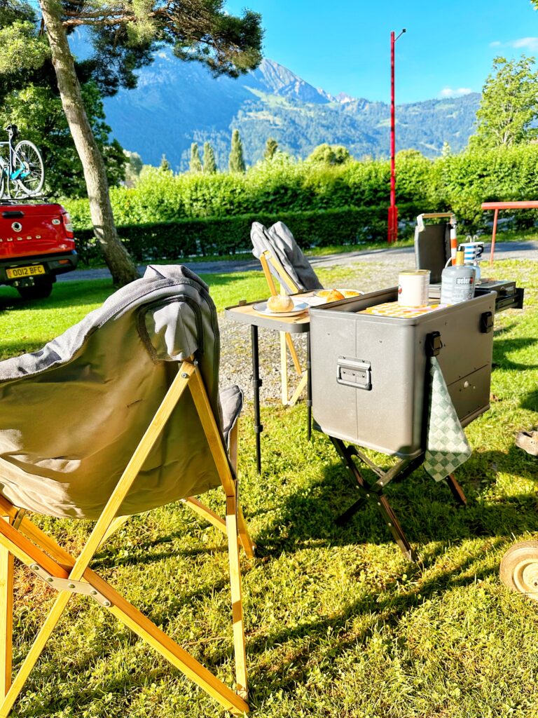 Campingküche Layzee und Stühle auf grüner Wiese mit spektakulärem Alpenpanorama und rotem Pickup im Hintergrund am Camping Alpenblick