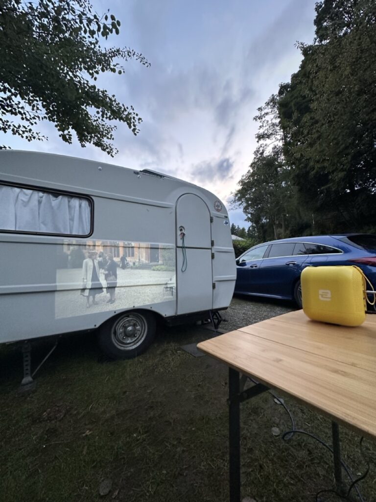 Weißer Wohnwagen auf Campingplatz bei Dämmerung, an dessen Seitenwand ein Film projiziert wird. Im Vordergrund ein Holztisch mit gelber EcoFlow Powerstation, dahinter ein blaues Auto zwischen Bäumen.