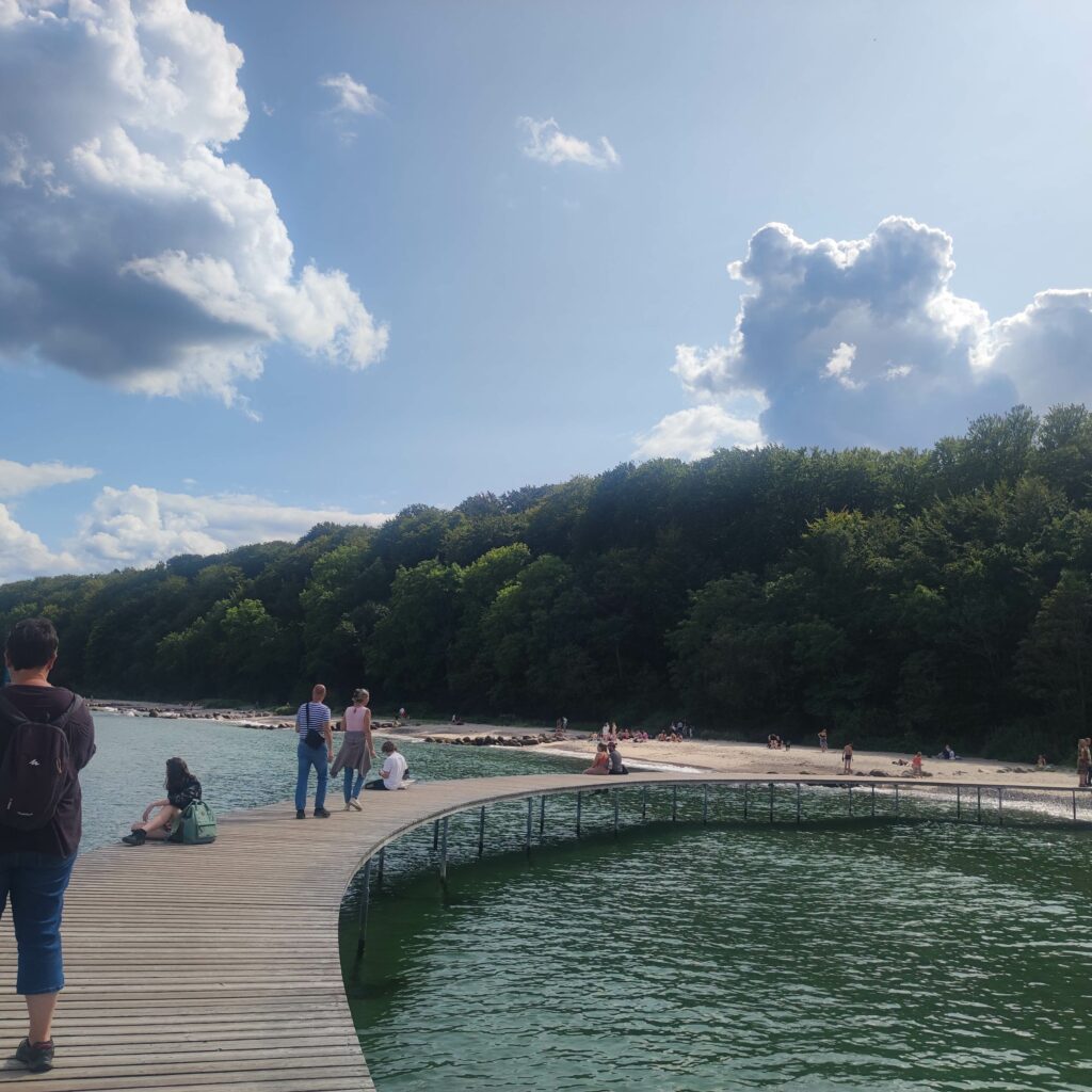 Holzsteg über Wasser Richtung bewaldeter Küste, Menschen entspannen am Ufer unter wolkigem Himmel.