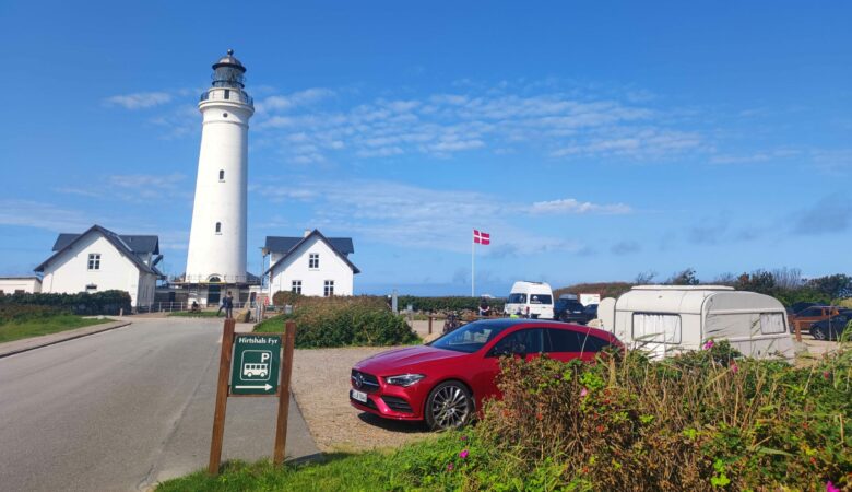 Weißer Leuchtturm in Hirtshals, Dänemark mit roten Auto, Wohnwagen und dänischer Flagge bei blauem Himmel.
