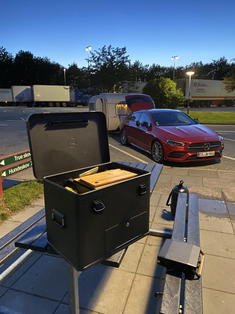 Geöffneter Campingkocher auf Rastplatz neben rotem Auto mit Wohnwagen, beleuchtet von Straßenlampen am Abend.