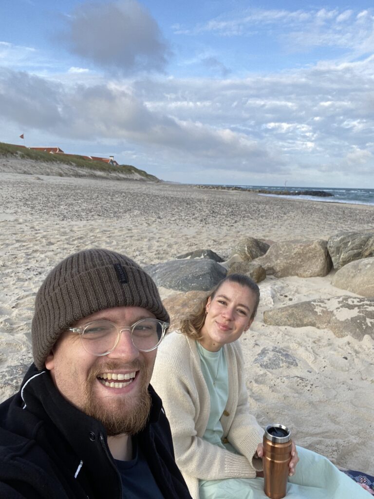 Zwei Personen sitzen am Sandstrand vor großen Steinen, dahinter Dünen, Häuser und das Meer unter wolkigem Himmel.