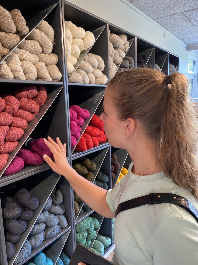 Person betrachtet und berührt bunte Wollknäuel in einem Schaufenster mit Regalen voller sorgfältig sortierter Wolle.
