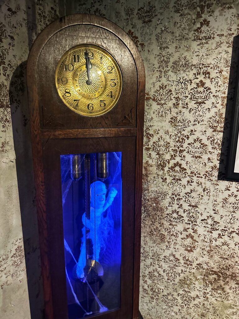 Eine altehrwürdige Standuhr mit einem goldenen Zifferblatt, die in einer der Ausstellungen des Augsburger Puppenkiste Museums "Die Kiste" steht. Im Inneren des Pendelbereichs ist eine hellblau leuchtende, geisterhafte Figur in Spinnweben gehüllt zu sehen, was der Uhr ein mystisches und leicht gruseliges Aussehen verleiht.