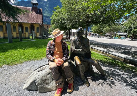 Phil sitzt neben der Bronze-Statue von Sherlock Holmes vor dem Museum in Meiringen mit der ehemaligen Kirche im Hintergrund