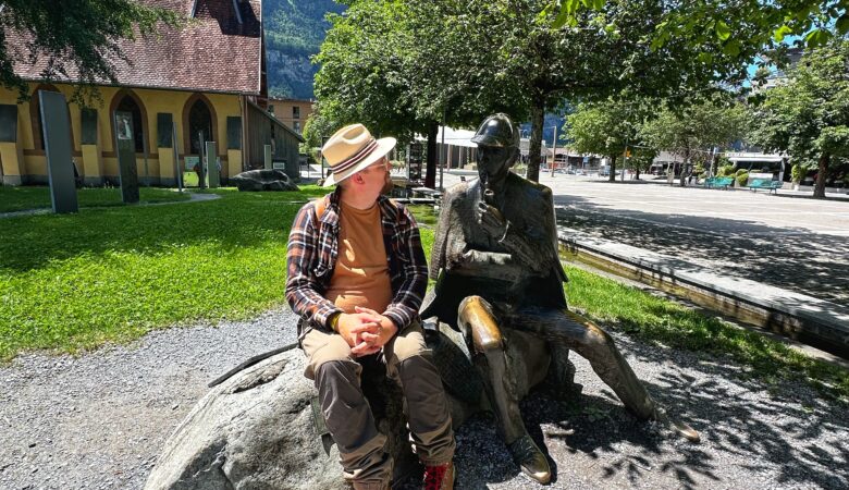 Phil sitzt neben der Bronze-Statue von Sherlock Holmes vor dem Museum in Meiringen mit der ehemaligen Kirche im Hintergrund