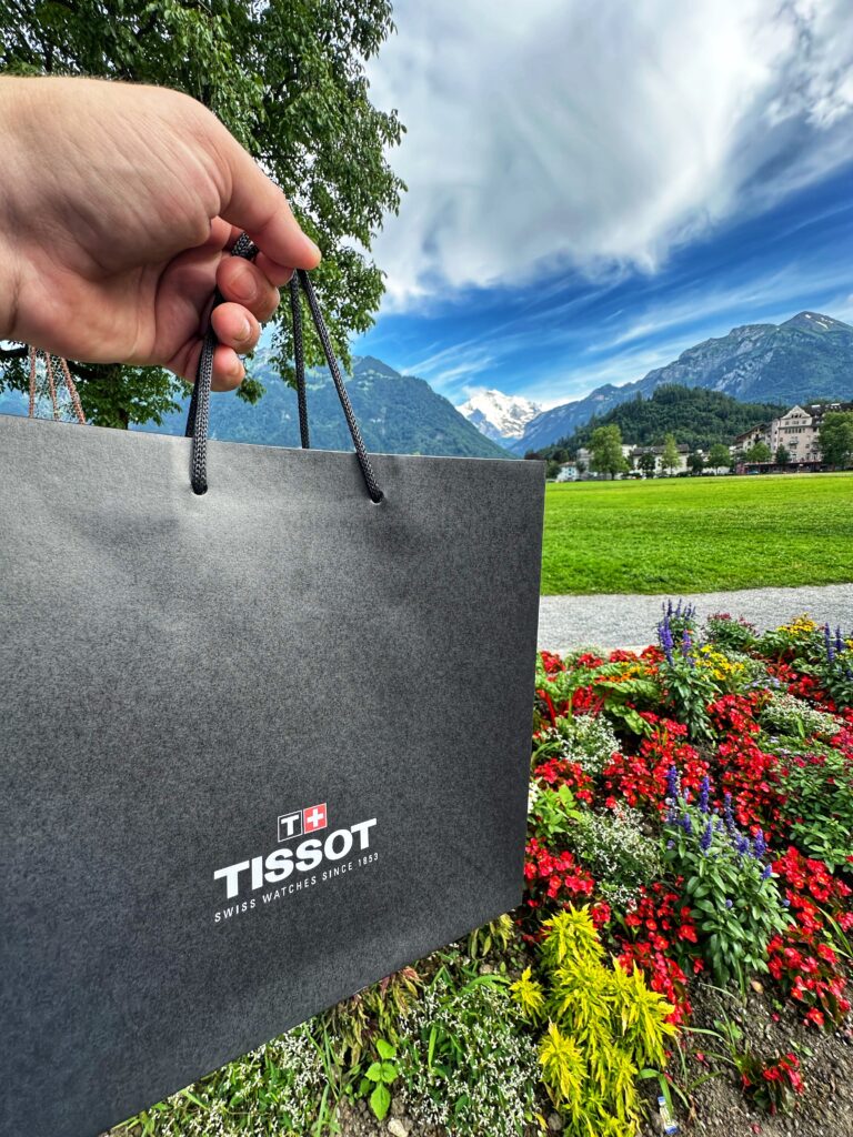 Hand hält graue Tissot Einkaufstasche vor Schweizer Bergkulisse mit bunten Blumen und Jungfrau-Massiv