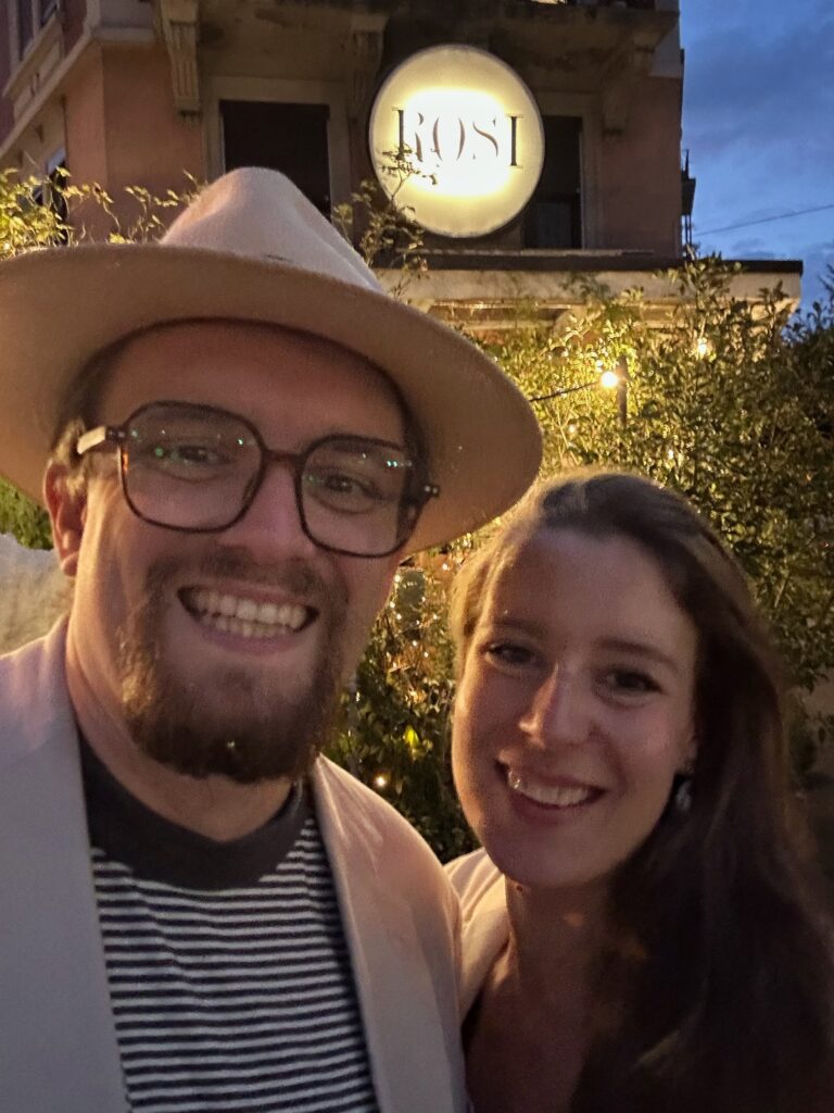 Phil und Anna lächeln glücklich vor dem beleuchteten ROSI Restaurant in Zürich bei Dämmerung