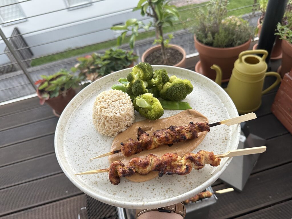 Yakitori-Spieße mit Erdnusssoße, Vollkornreis und gedämpftem Gemüse auf einem Teller vor Balkonpflanzen