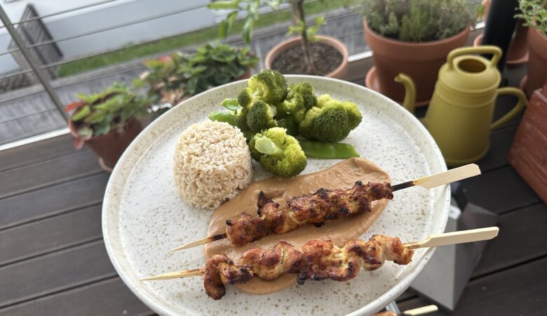 Yakitori-Spieße mit Erdnusssoße, Vollkornreis und gedämpftem Gemüse auf einem Teller vor Balkonpflanzen