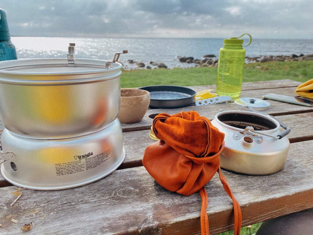 Ein rustikaler Holztisch an einer Küste mit Outdoor-Ausrüstung. Ein Trangia-Kocher steht links, daneben ein geöffneter orangefarbener Lederbeutel und die Trangia-Kaffeekanne, in die bereits frisches Kaffeepulver gefüllt wurde. Im Hintergrund sieht man eine Wiese, steinige Küste, das Meer und eine grüne Nalgene-Trinkflasche.