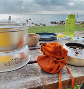 Ein rustikaler Holztisch an einer Küste mit Outdoor-Ausrüstung. Ein Trangia-Kocher steht links, daneben ein geöffneter orangefarbener Lederbeutel und die Trangia-Kaffeekanne, in die bereits frisches Kaffeepulver gefüllt wurde. Im Hintergrund sieht man eine Wiese, steinige Küste, das Meer und eine grüne Nalgene-Trinkflasche.