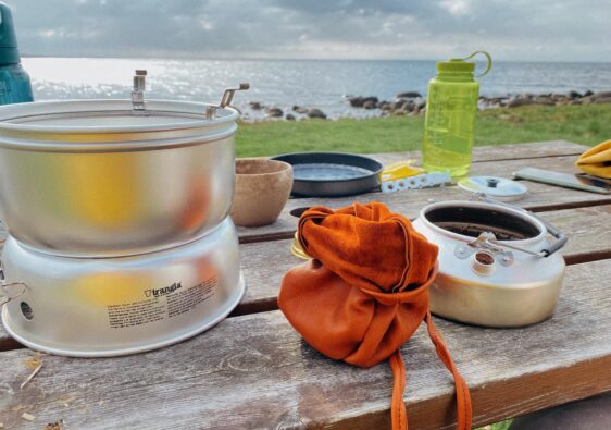 Ein rustikaler Holztisch an einer Küste mit Outdoor-Ausrüstung. Ein Trangia-Kocher steht links, daneben ein geöffneter orangefarbener Lederbeutel und die Trangia-Kaffeekanne, in die bereits frisches Kaffeepulver gefüllt wurde. Im Hintergrund sieht man eine Wiese, steinige Küste, das Meer und eine grüne Nalgene-Trinkflasche.