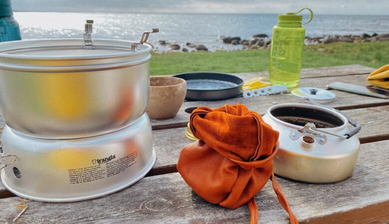 Ein rustikaler Holztisch an einer Küste mit Outdoor-Ausrüstung. Ein Trangia-Kocher steht links, daneben ein geöffneter orangefarbener Lederbeutel und die Trangia-Kaffeekanne, in die bereits frisches Kaffeepulver gefüllt wurde. Im Hintergrund sieht man eine Wiese, steinige Küste, das Meer und eine grüne Nalgene-Trinkflasche.