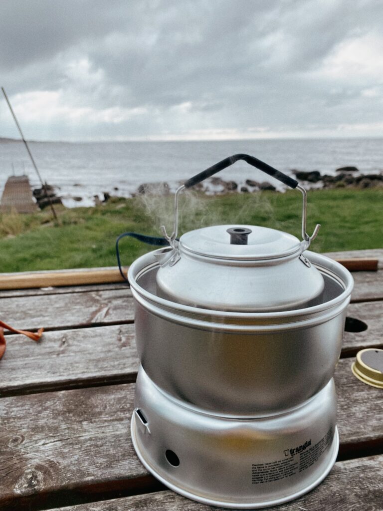 Nahaufnahme eines voll aufgebauten Trangia-Sturmkochers auf einem Holztisch. Aus der kleinen Aluminium-Kaffeekanne auf dem brennenden Kocher steigt heißer Dampf auf. Im Hintergrund ist eine steinige Küste und das Meer unter einem leicht bewölkten Himmel zu sehen.
