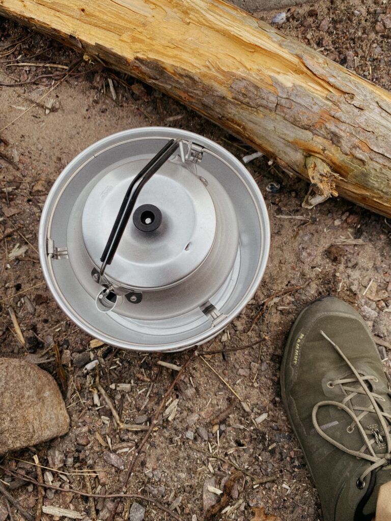 Ein Trangia Sturmkocher aus Aluminium mit eingesetzter Kaffeekanne steht auf steinigem Waldboden direkt neben einem umgestürzten Baumstamm. Rechts im Bild ist ein Fuß in einem olivgrünen Mammut-Wanderschuh zu sehen.