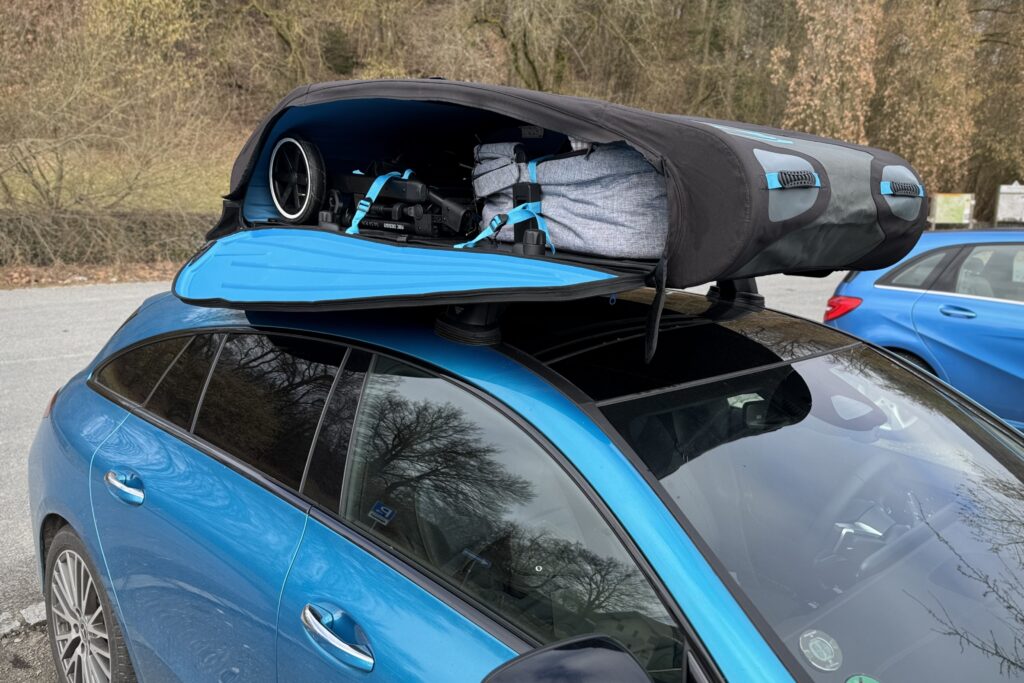 Ein blauer Mercedes CLA Shooting Brake mit einer geöffneten, faltbaren Dachbox (Norauto Bermude Flex) auf dem Dach. Im Inneren der schwarzen Dachbox mit blauen Akzenten sind ein zusammengeklappter Kinderwagen und eine graue Reisetasche sicher mit blauen Spanngurten verzurrt.