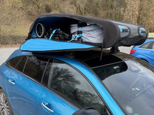Ein blauer Mercedes CLA Shooting Brake mit einer geöffneten, faltbaren Dachbox (Norauto Bermude Flex) auf dem Dach. Im Inneren der schwarzen Dachbox mit blauen Akzenten sind ein zusammengeklappter Kinderwagen und eine graue Reisetasche sicher mit blauen Spanngurten verzurrt.