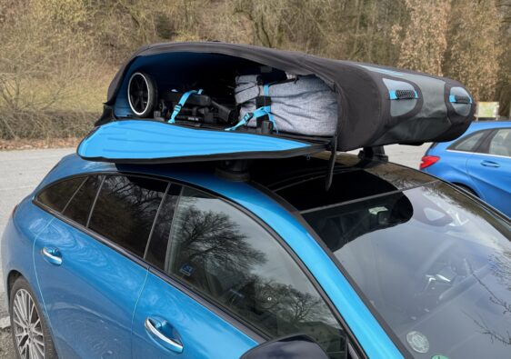 Ein blauer Mercedes CLA Shooting Brake mit einer geöffneten, faltbaren Dachbox (Norauto Bermude Flex) auf dem Dach. Im Inneren der schwarzen Dachbox mit blauen Akzenten sind ein zusammengeklappter Kinderwagen und eine graue Reisetasche sicher mit blauen Spanngurten verzurrt.
