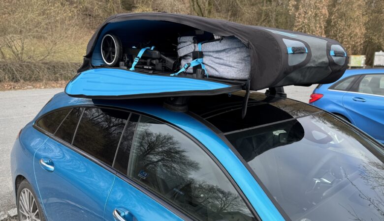 Ein blauer Mercedes CLA Shooting Brake mit einer geöffneten, faltbaren Dachbox (Norauto Bermude Flex) auf dem Dach. Im Inneren der schwarzen Dachbox mit blauen Akzenten sind ein zusammengeklappter Kinderwagen und eine graue Reisetasche sicher mit blauen Spanngurten verzurrt.