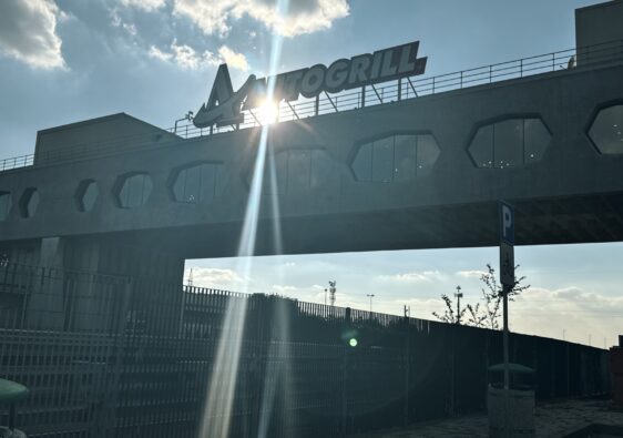 Aussicht auf das architektonische Brückenrestaurant Autogrill Limenella über der Autobahn, mit strahlendem Sonnenlicht und dem "AUTOGRILL"-Schriftzug.
